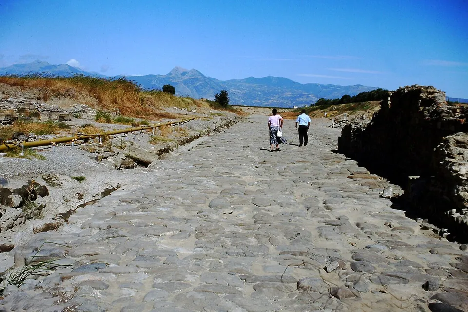Two people walking along a broad street paved with large worn stones, a ruined wall to the right, dry grass to the left and a mountain range on the horizon under a blue sky.