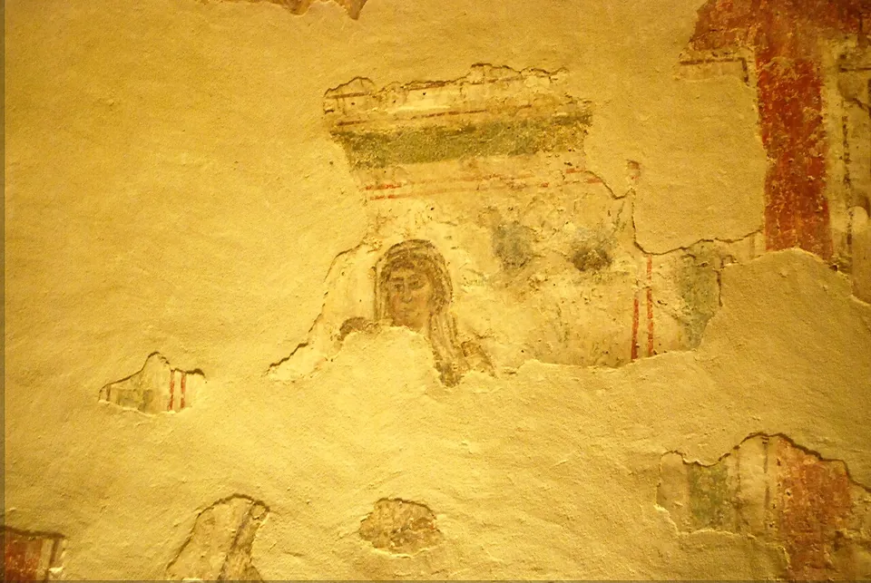 A fragment of wall painting on yellowish plaster, showing the head and shoulders of a woman framed by painted architecture, with patches of red and green around it.