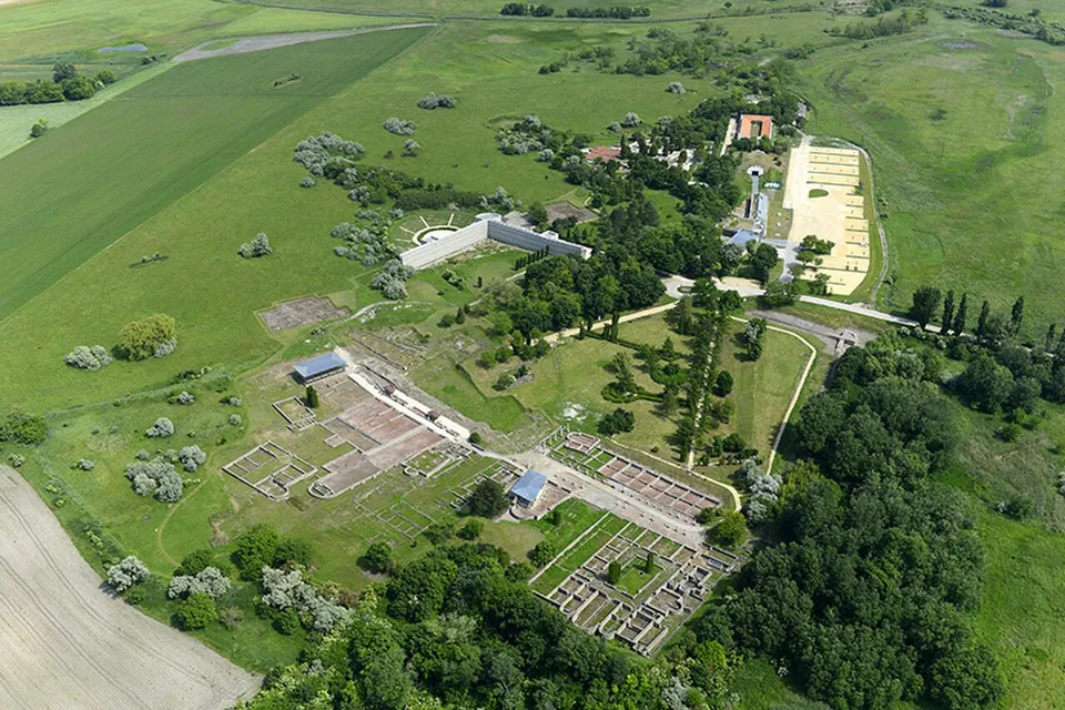 An aerial view of the ruin field among fields and woodland, with grids of excavated foundations, a straight tree-lined avenue, white modern buildings and a large pale rectangle of ground beyond.