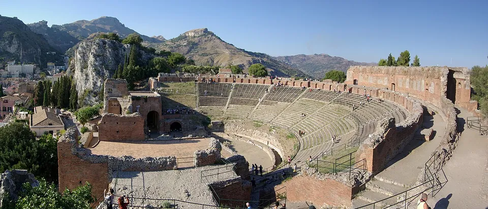 A panorama of the whole theatre from high on one side of the seating bowl, with the rows of the seating bowl sweeping round to a ruined red-brick stage building on the left, and the houses of the town and steep rocky mountains behind.