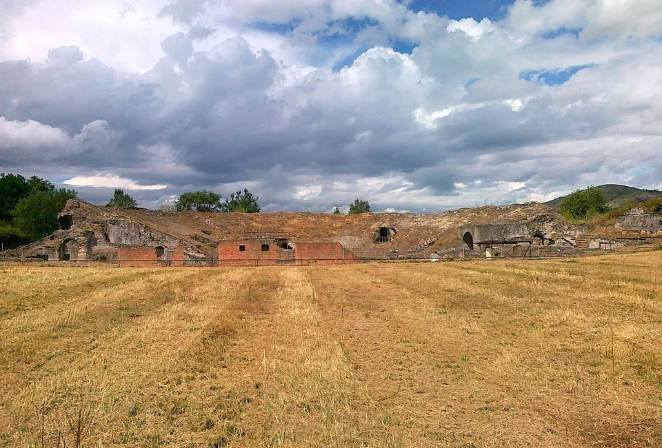A long, low ruin of rubble masonry with a row of dark arched openings and patches of red brick, seen across a wide field of mown, straw-coloured grass under a cloudy sky.