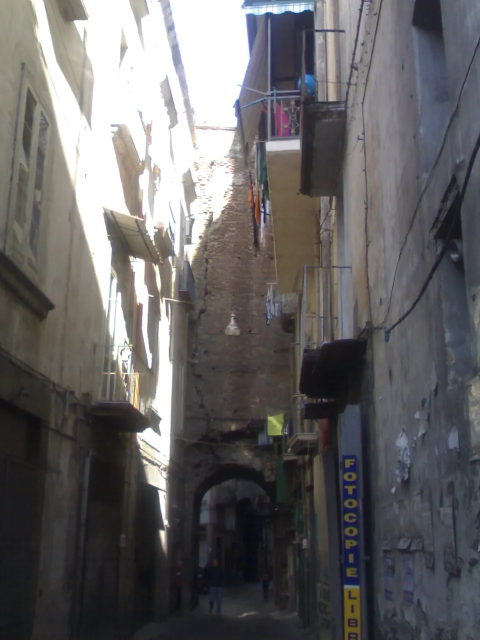 A narrow street between tall plastered buildings with shuttered windows and laundry, spanned in the middle distance by a high round arch of brick.