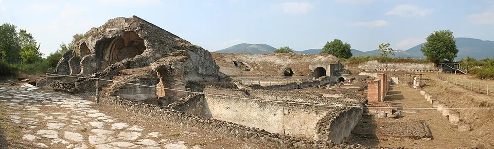 A panorama of the excavated theatre, with a paved street of large polygonal stones in front, the high rubble mass of the seating behind it, and the low walls of the stage building stretching to the right, with hills on the horizon.