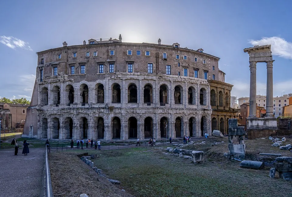 The curved outer wall of the theatre, two storeys of arches in weathered travertine framed by half-columns, with a plainer palazzo with rows of windows built on top; in the foreground a lawn scattered with stone blocks, and at the right tall marble columns carrying a piece of entablature.