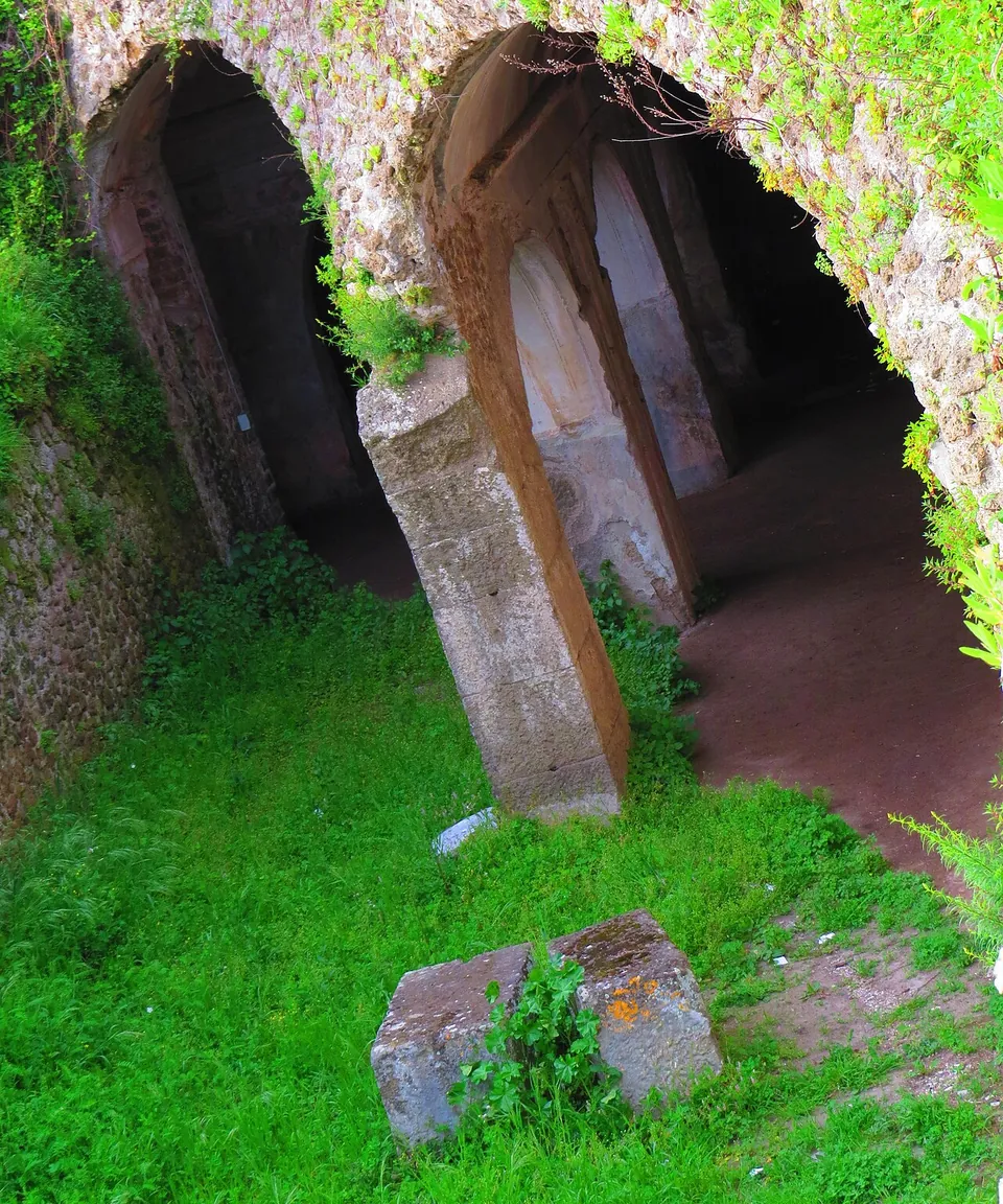 Looking down at two arched openings in a rubble-stone wall, separated by a square pier of cut stone, with pale plastered pilasters and a dark vaulted corridor inside and grass in the foreground.