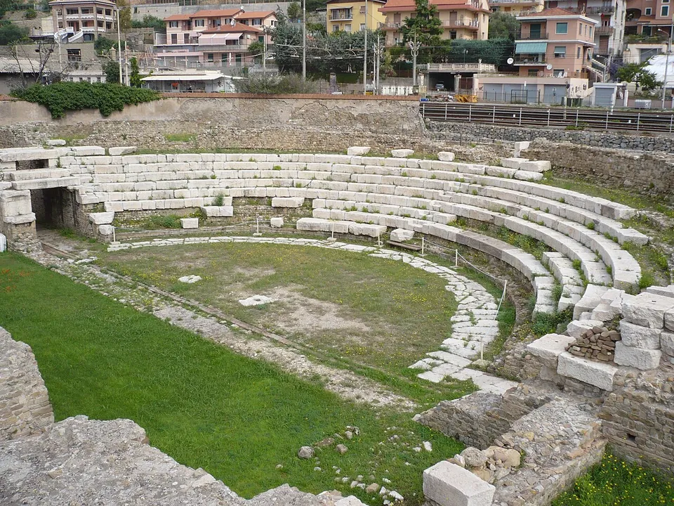 A small Roman theatre seen from one side, its curved rows of pale stone seats rising around a grassy orchestra edged with a paved strip, with apartment blocks beyond the outer wall.