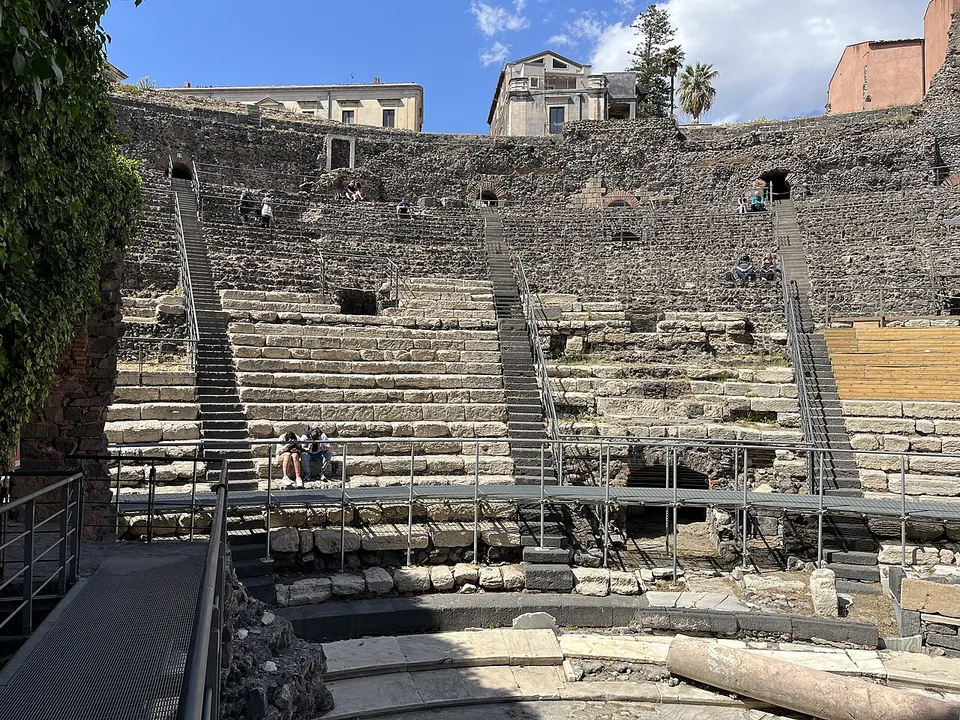 Tiers of grey stone seats rising in a semicircle, crossed by metal stairways and a raised walkway, with a few visitors sitting on the steps and buildings along the top.