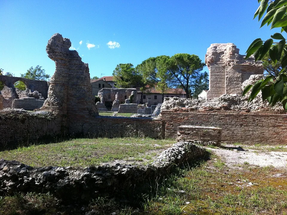 Brick walls and two broken masonry piers standing a few metres high under a blue sky, with a curved line of rubble foundations in the foreground and modern houses behind.