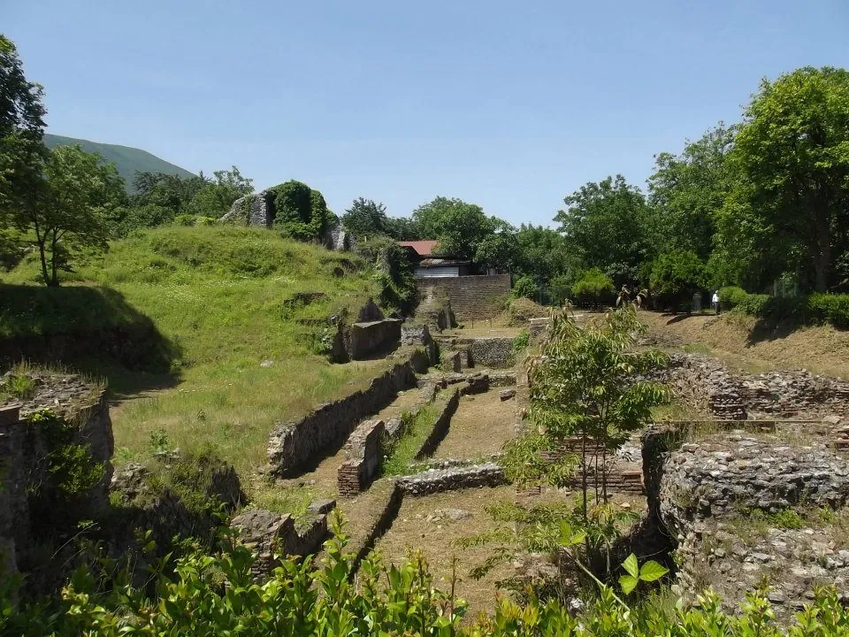Low walls of stone and brick stepping down a grassy slope among trees, with a house and wooded hills behind.