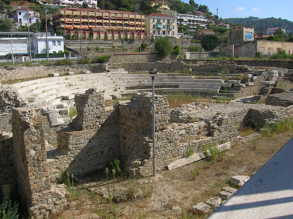 Ruined rubble-stone walls of different heights in the foreground, the curve of the theatre's stone seating beyond them, and the apartment blocks and houses of the modern town on the hillside behind.