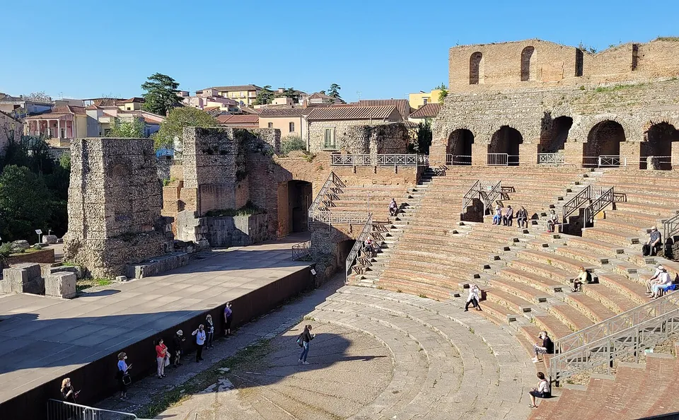 The whole theatre from beside the stage, the semicircle of seating with a few visitors on it to the right, the paved stage area and tall ruined walls beyond it to the left, and the houses of the old town behind.
