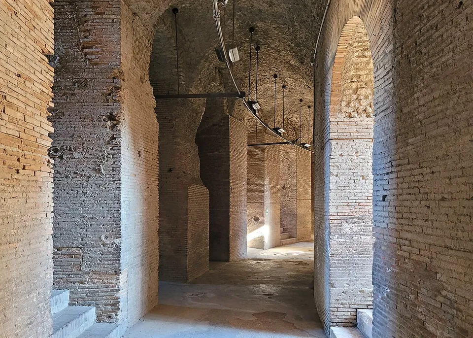 A vaulted passage of brick, lit by a row of hanging lamps, opening through an arch into further corridors with daylight at the far end.