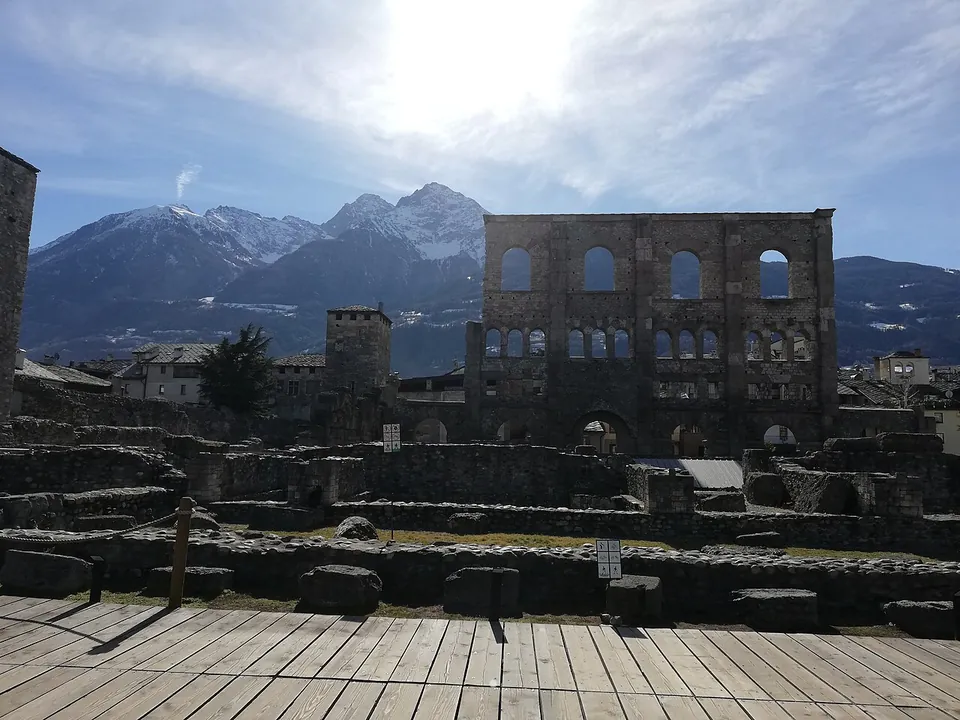 Seen from a wooden walkway, rows of low excavated stone walls and scattered blocks in front of the tall arched wall of the theatre, with the town's roofs and towers around it and snow-capped mountains behind.
