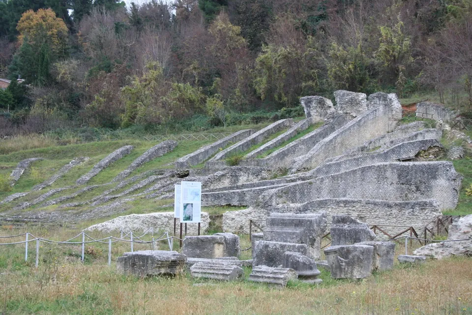 Stepped stone walls of the seating rise up a grassy slope, with loose carved blocks and an information panel in front and bare trees on the hillside behind.