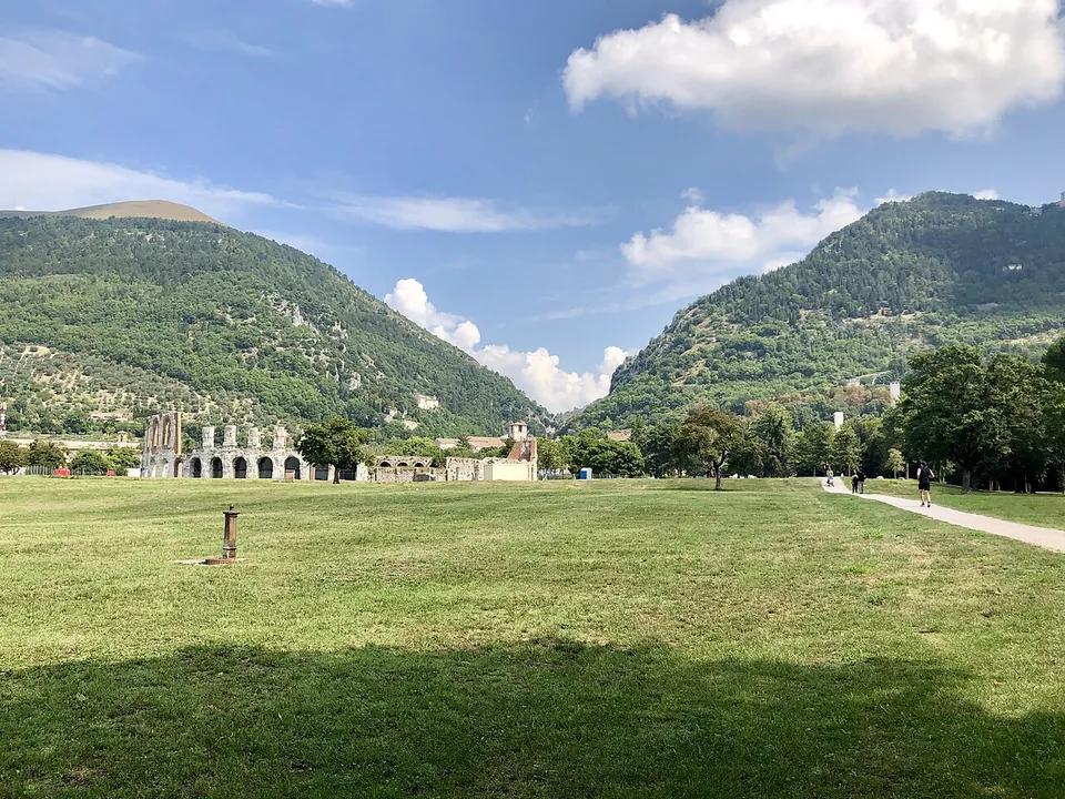 A wide lawn with a footpath crossing it towards a low line of pale stone arches in the distance, with wooded mountains rising steeply behind.