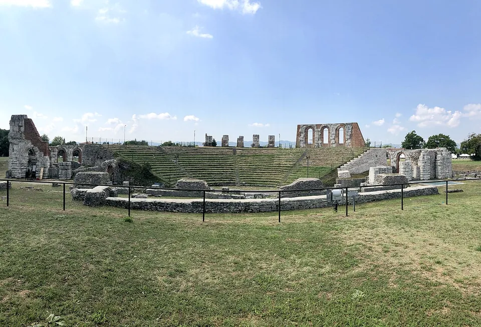 The theatre seen from the stage side - the low stone foundations of the stage behind a metal railing in the foreground, the tiers of seating beyond, and the standing arches at the top of the curve.