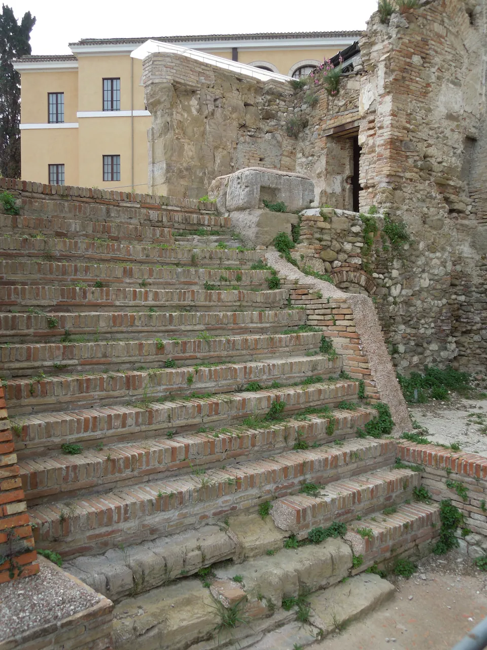 A broad flight of steps edged in brick climbing between rough stone walls inside the excavated theatre, with a modern yellow building above.