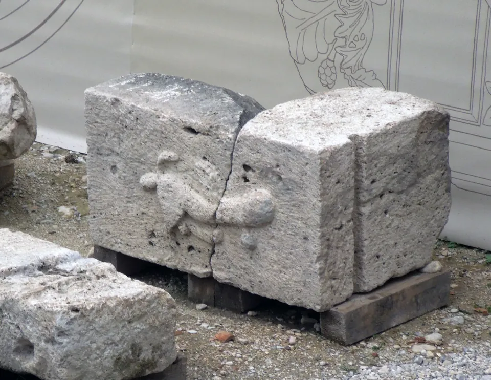 A pale, pitted travertine block, cracked in two, carved in low relief with a winged phallus, resting on wooden battens on gravel in front of a printed hoarding.