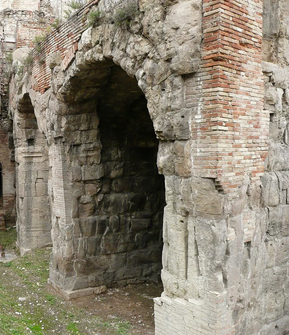 Two tall round-headed arches built of large grey squared stone blocks, patched and topped with red brick, standing in a sunken excavation with grass and earth at their feet.