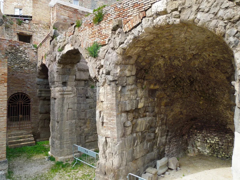 Massive piers of squared grey stone carrying round arches, with brick vaulting inside the nearer arch, a barred doorway in a brick wall behind, and a temporary metal barrier on the grass.