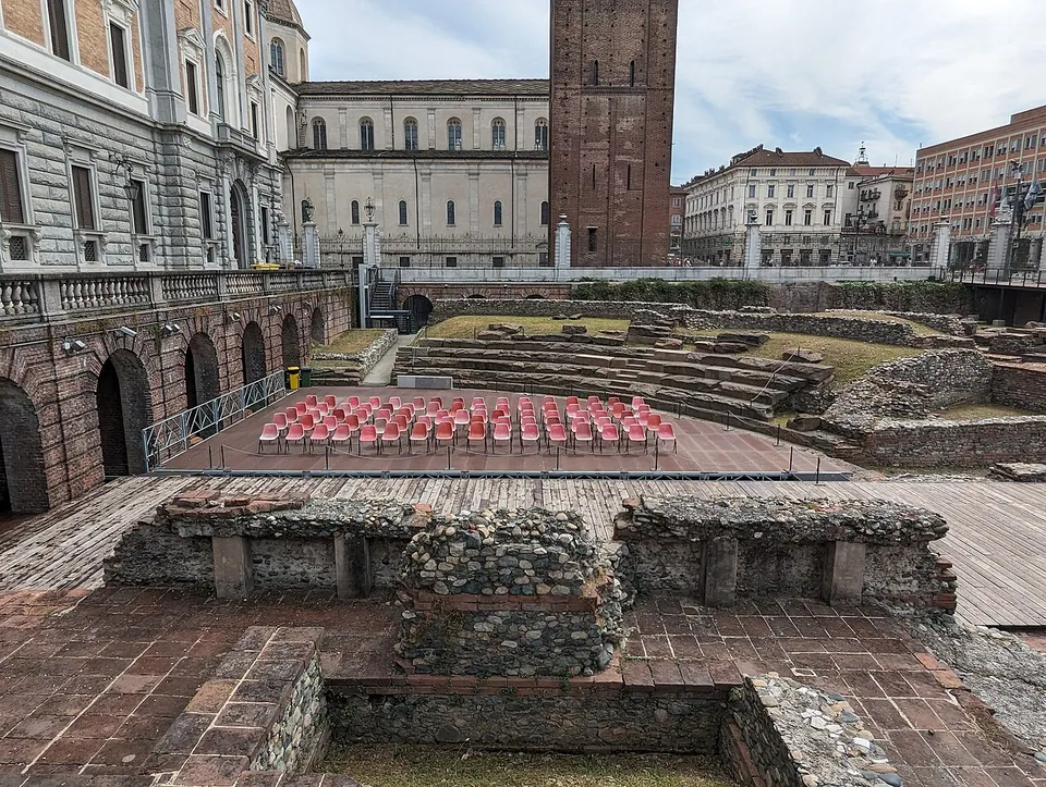 Excavated stone and brick foundations in the foreground, a paved stage area set with rows of red chairs, and grassed curved steps of the seating behind, enclosed by the arcaded wall of a palace wing with a tall brick bell tower beyond.