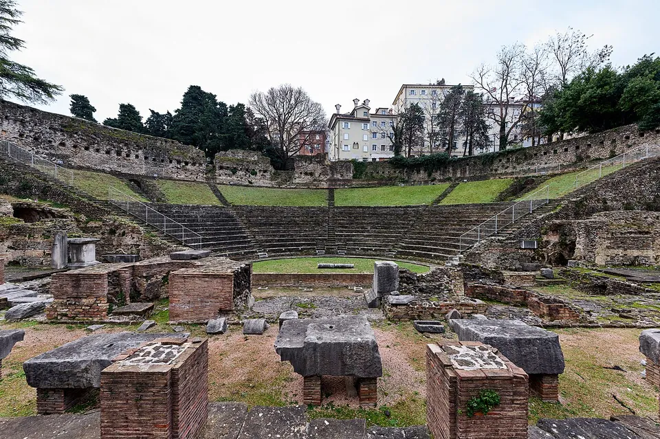 A wide view across the ruins toward the seating, which rises in grey stone rows to grass terraces and a long high wall, with trees and pale apartment blocks above it; brick piers and fallen stone blocks fill the foreground.