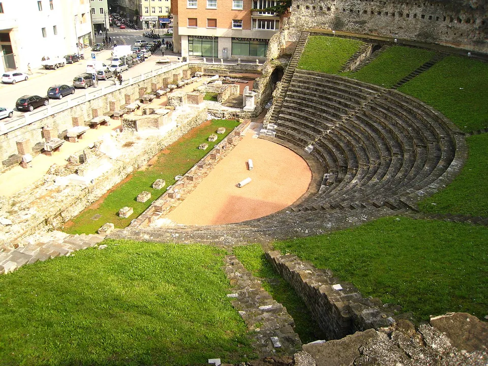 The Roman theatre seen from above, a half-circle of stone seating rows set into a grassy slope around a bare orchestra floor, with the low foundations of the stage building in front and a street with parked cars beyond.