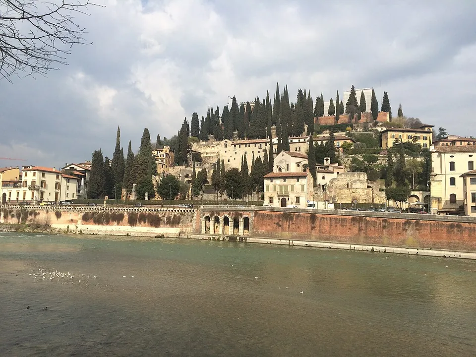 A steep hillside across a broad green river, covered in tall cypresses and pale houses, with a red-brick building on the summit and a high brick embankment wall with a row of small arches along the water.