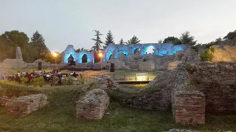 An audience seated on chairs at dusk in front of a lit stage, with the arches of the ruined theatre floodlit in blue behind them and brick ruins in the foreground.