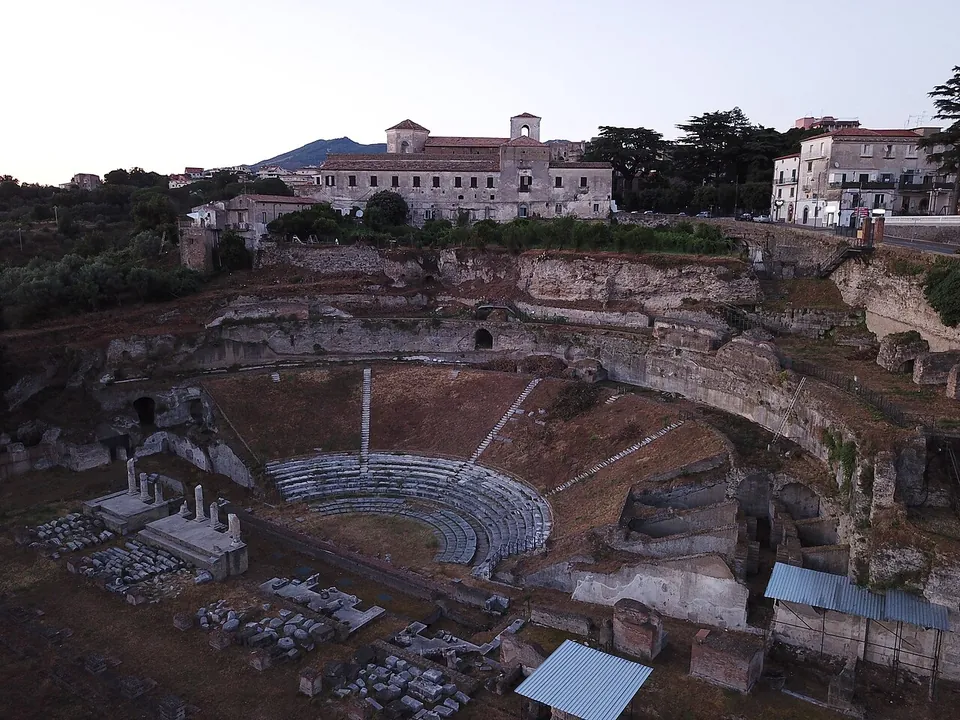 An aerial view at dusk of a large semicircular theatre cut into a hillside, its lower rows of stone seating visible, with the stage area of scattered column bases and stone blocks in front and a long multi-storey building on the ridge above.