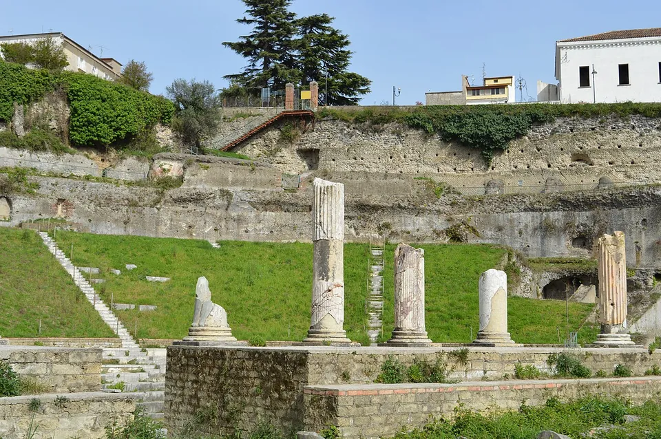 Five marble column shafts of different heights standing on a stone platform, one of them only a broken stump, with a grassy slope, stone stairs and high rubble retaining walls behind.