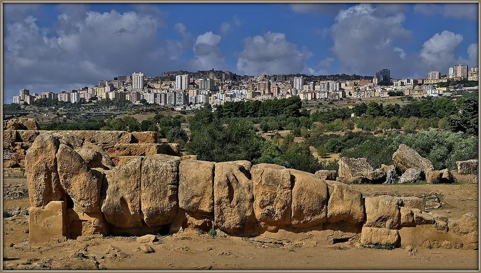 A colossal human figure assembled from stacked stone blocks, lying on its back on the ground, with the apartment blocks of modern Agrigento on the hill behind.