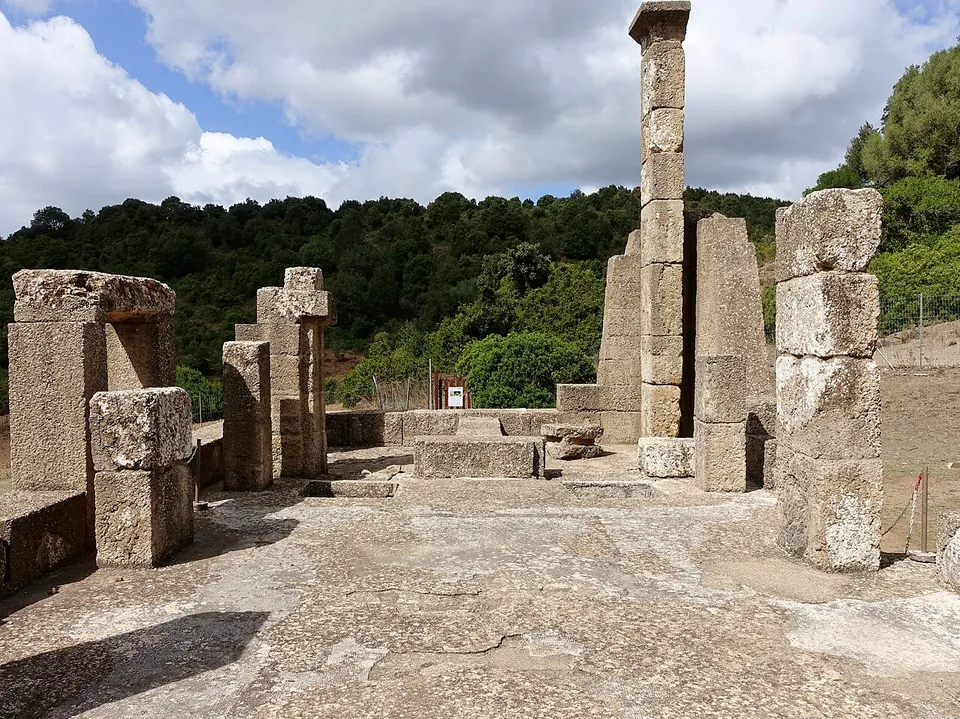 Standing on the temple platform among squared limestone blocks and pillars of uneven height, with a smooth worn floor in front, one tall shaft rising in the middle and a wooded hillside beyond.