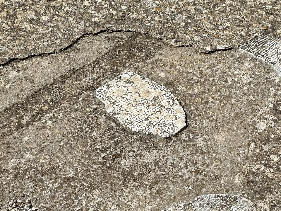 A close-up of a worn grey floor with a patch of small white tesserae edged in dark ones set into it, and another strip of mosaic at the edge of the frame.