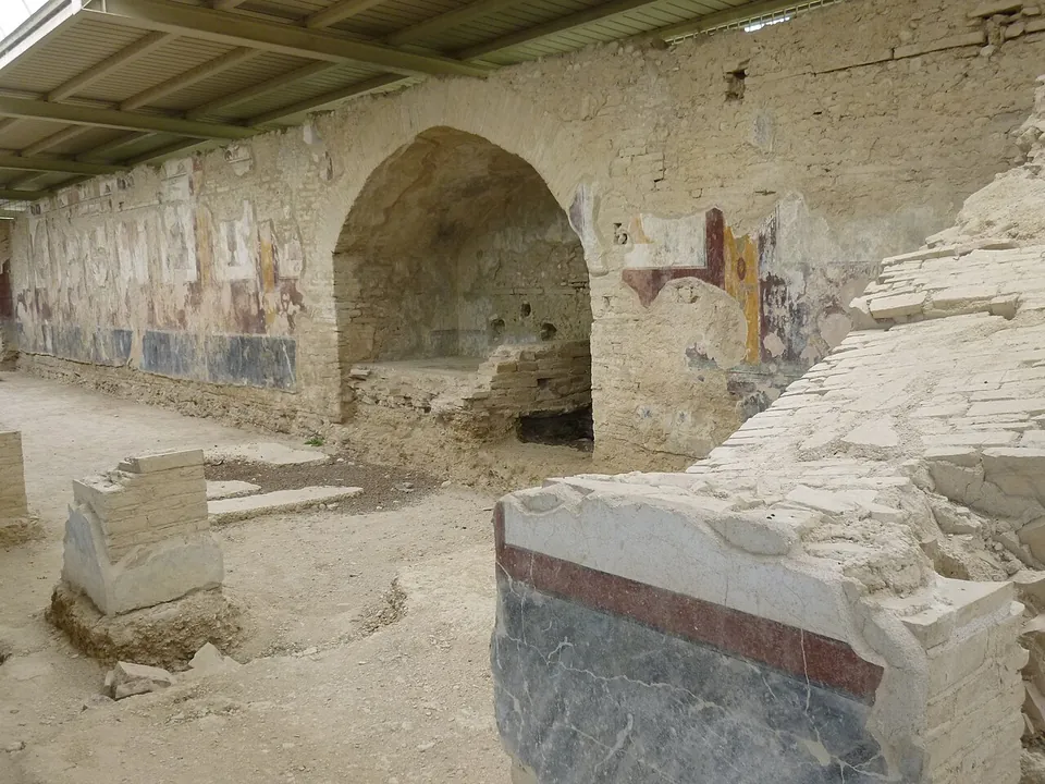 A long sunken gallery under a modern roof of metal and translucent panels, its stone wall painted with faded red, yellow and white panels above a dark blue base, a metal walkway running along it and a few visitors at the far end.