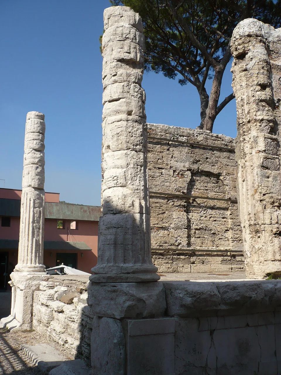 Two weathered stone columns without capitals stand at the front of a temple podium, before a wall of squared stone blocks, with a pine tree and a pink house behind.