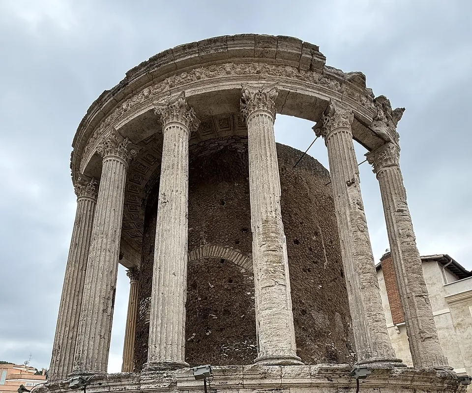 A round temple seen from below, its curved row of fluted Corinthian columns carrying a carved frieze and cornice in front of a rough circular inner wall, against a cloudy sky.