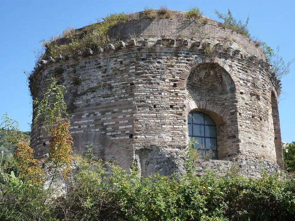 A round building of brick banded with stone, with an arched window and a row of projecting stone brackets below its domed top, rising above green bushes against a blue sky.