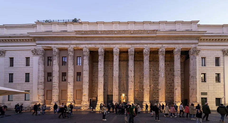 A row of tall fluted columns with Corinthian capitals standing in front of a dark, pitted stone wall, set into the middle of a pale later building, with people walking in the square in front at dusk.