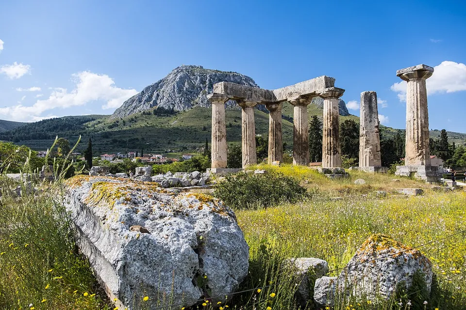 Seven weathered monolithic columns of a Doric temple, still carrying a stretch of their beam, on a grassy rise strewn with stone blocks and yellow flowers, with the great rock of Acrocorinth behind.