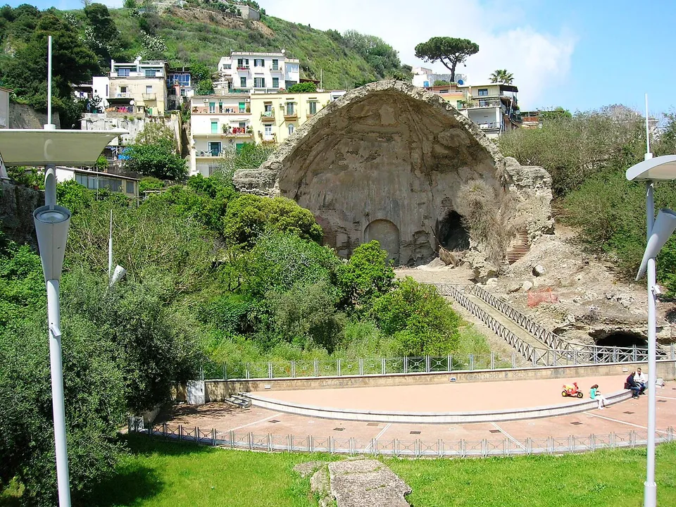 A massive ruined vault of stone, broken open down one side, standing on a green slope behind railings and a paved semicircular terrace, with apartment blocks on the hill above it.