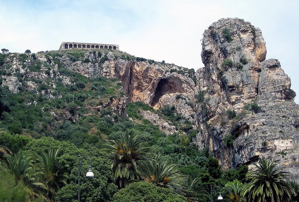 A scrubby green hillside rising to pale limestone cliffs with a deep hollow in the rock face and a separate rock pinnacle on the right, a long arcaded wall running along the skyline, and palm trees and a street lamp in the foreground.