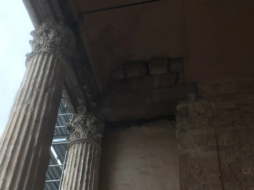 Looking up between two fluted columns to their carved Corinthian capitals and the stone beams of the entablature they carry, with the back wall of the porch in shadow.