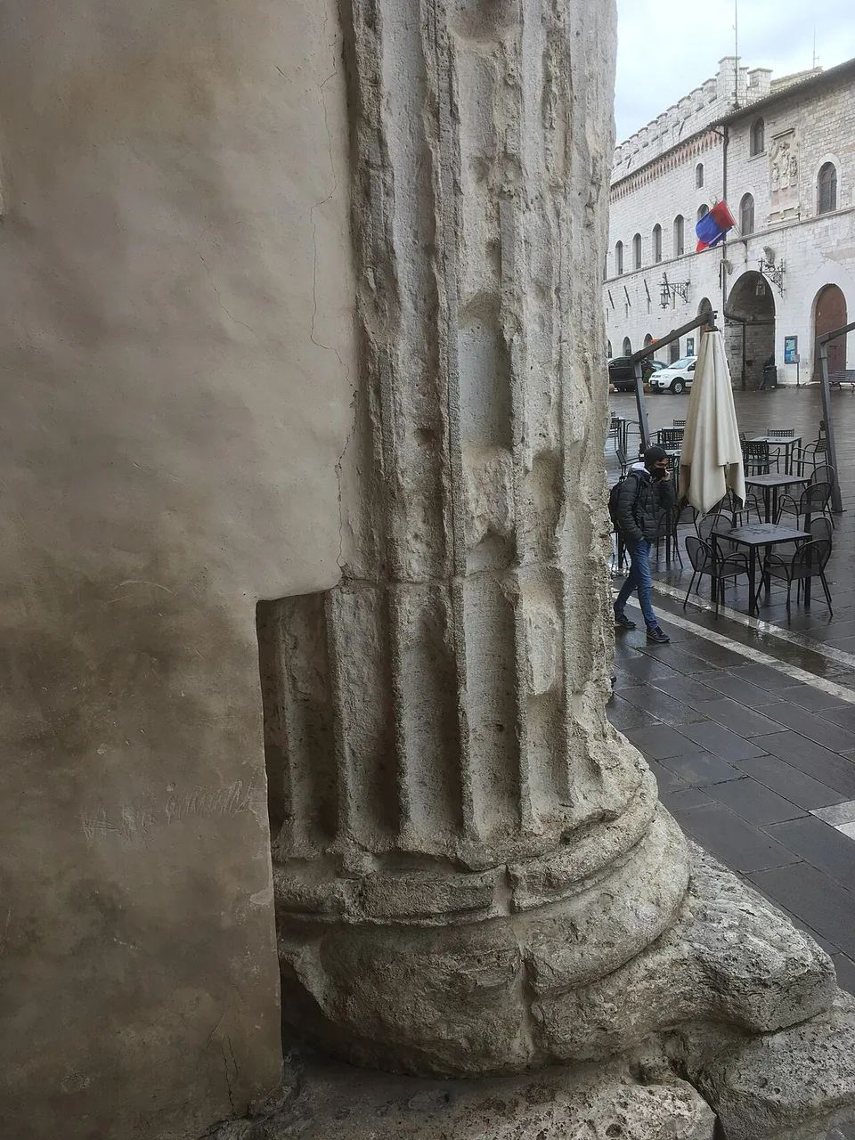 A close view of the foot of one column, its fluted shaft worn and chipped, resting on a moulded base on a square stone block, with the wet paving of the square and café tables beyond.