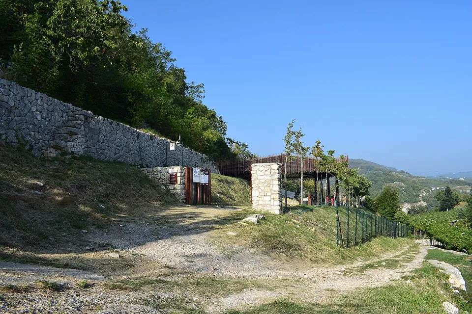 A gravel track running between a dry-stone terrace wall and a fence, past a small gated entrance and a stone pillar, towards the slatted roof of the shelter, with vineyard hills in the distance.