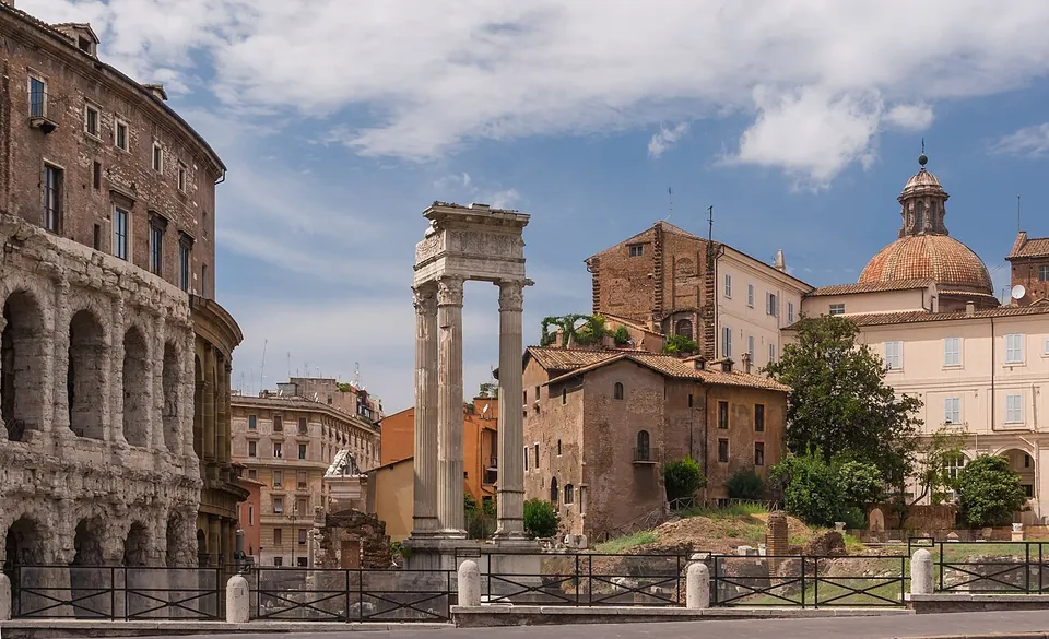 Three fluted white marble columns with Corinthian capitals standing on a podium and carrying a section of carved entablature, with the edge of the theatre's arcades at the left and a domed church behind.