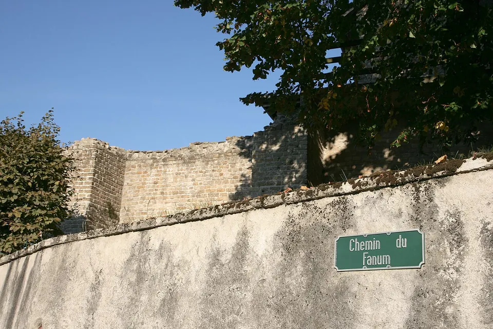 A tall wall of small squared stones rising behind a plastered retaining wall that carries a green street sign reading "Chemin du Fanum", with a tree overhanging.