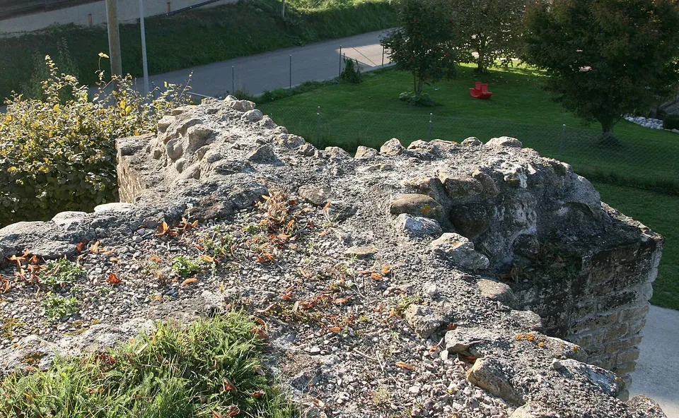 The weathered top of a thick wall of mortared rubble faced with squared stones, with a lawn and a road below.