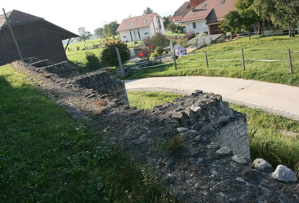 The top of a long low wall of rubble and squared stones running along grass beside a paved lane, with houses and a barn behind.