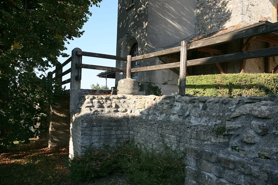 A stone wall of small squared blocks with a wooden railing on top, in front of the stone tower of a church with an arched window.